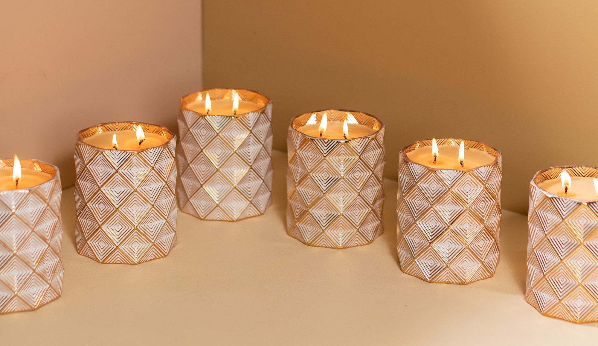 Six lit candles in patterned, white and gold holders are arranged on a beige surface against a two-tone cream and gold wall. The candles create a warm and cozy ambiance.