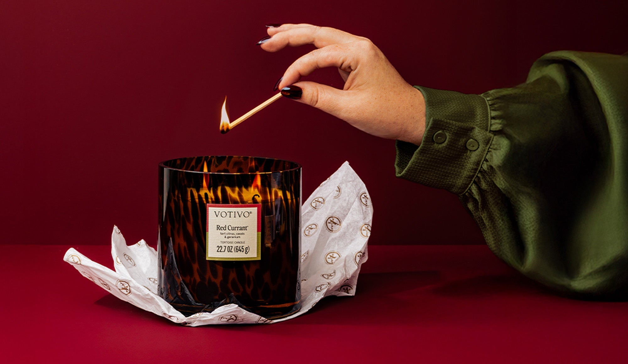 A hand in a green satin sleeve lights a Votivo Red Currant candle with a match. The candle, in a patterned glass jar, sits on white tissue paper against a deep red background.
