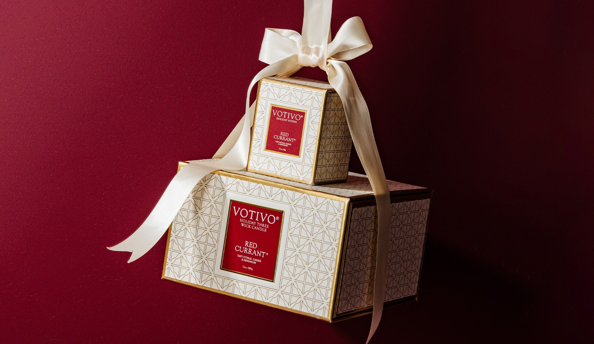 Two elegant cream and gold gift boxes with red labels reading Votivo Red Currant, stacked on top of each other and tied with a large ivory ribbon, against a dark red background.