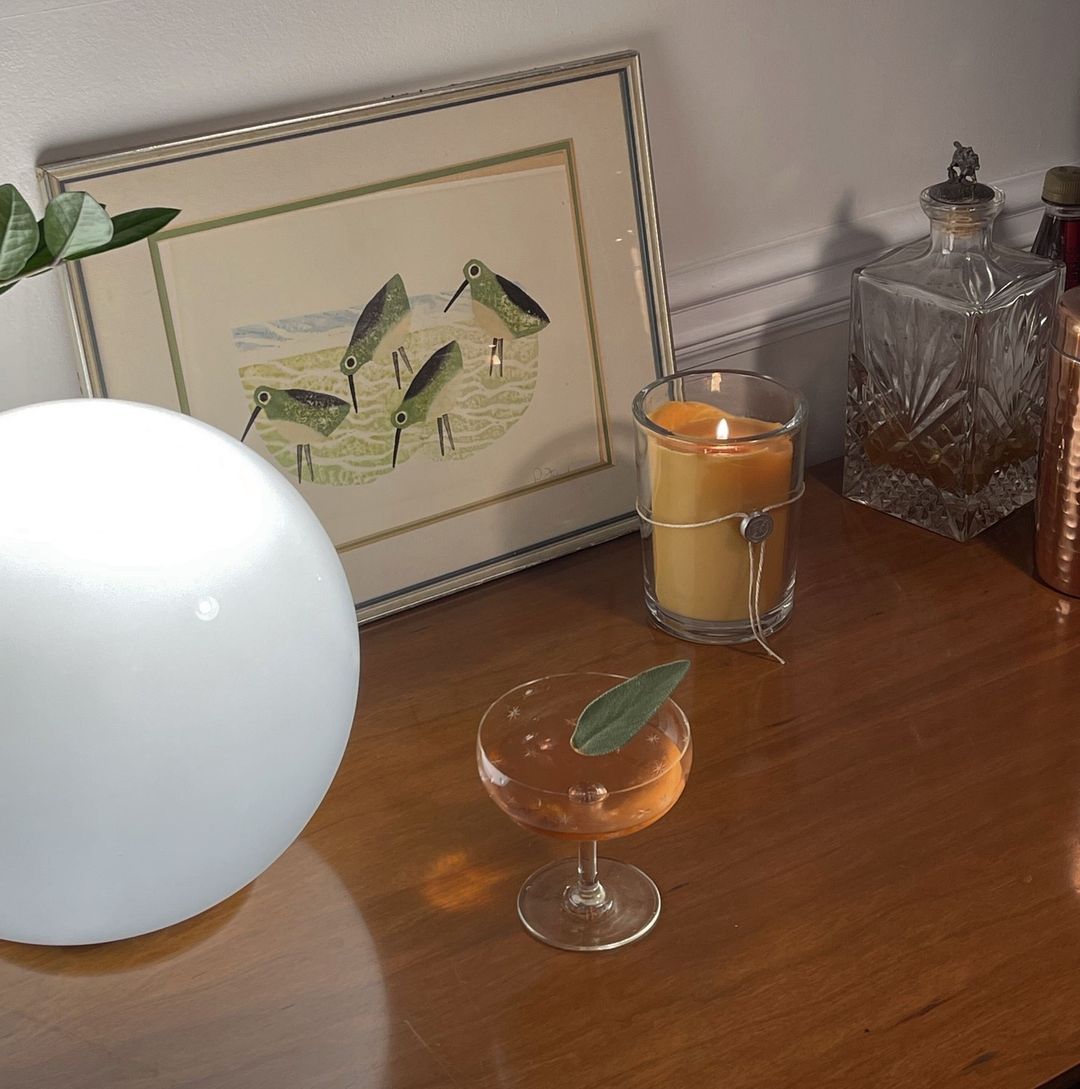 A cocktail garnished with a leaf sits on a wooden table next to a glowing orb lamp, framed bird art, the Votivo Honeysuckle 6.8 oz. Aromatic Candle, glass decanters, and a copper shaker.