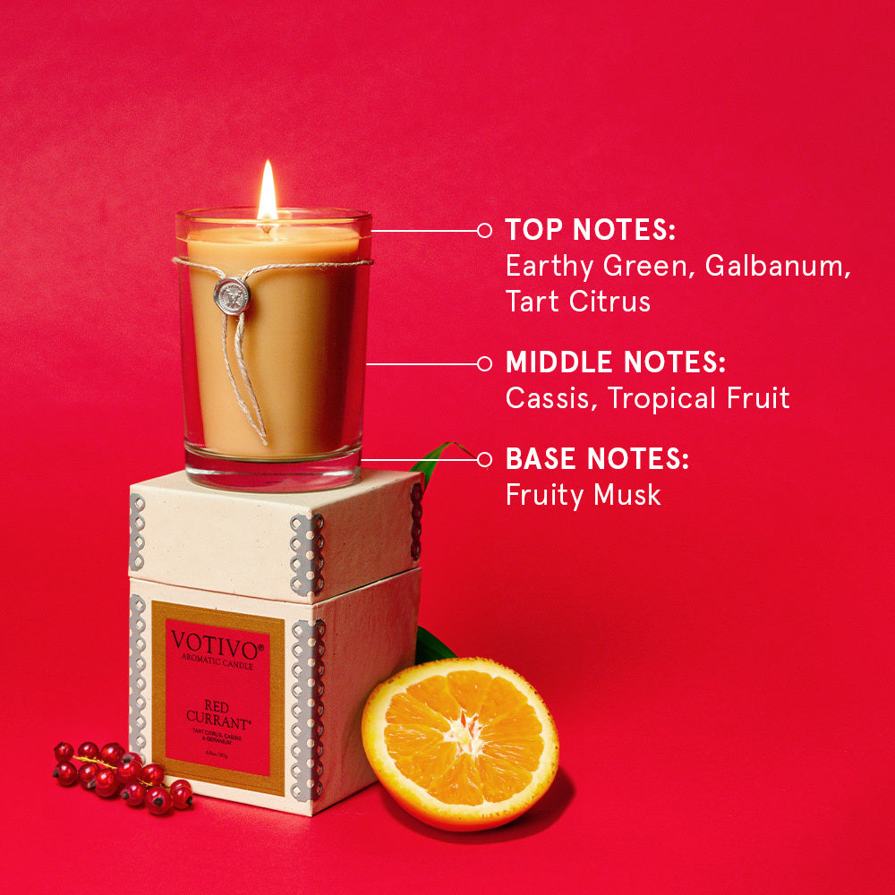 A Votivo Red Currant 6.8 oz. Aromatic Candle rests on its box with currants and orange slices nearby. The soy blend features earthy green, galbanum, tart citrus, cassis, tropical fruit, and fruity musk notes.