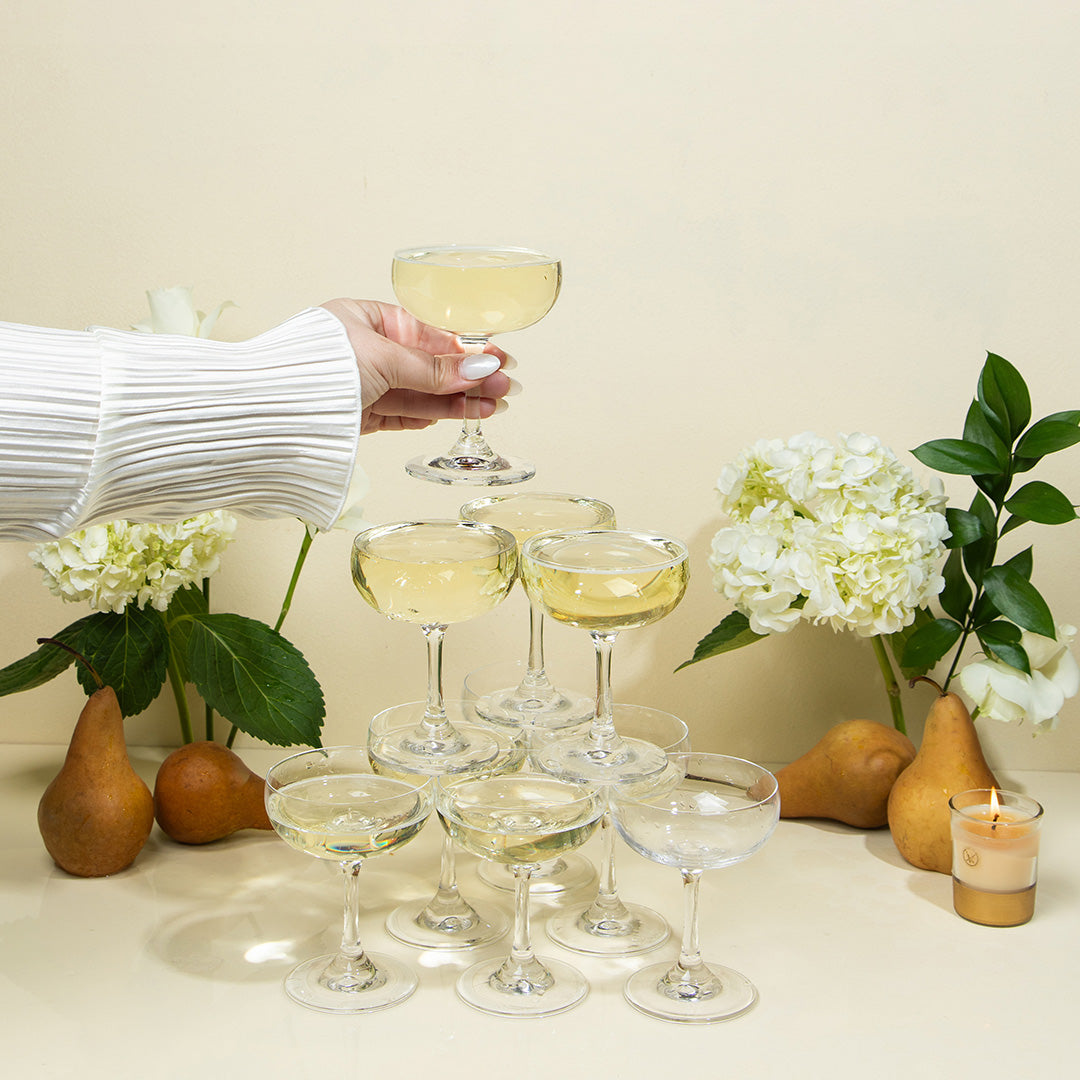 A hand in a white pleated sleeve holds a coupe glass atop a pyramid of filled coupe glasses, surrounded by pears, white hydrangeas, greenery, and the Votivo Gilded Pear 10 oz. Holiday Candle against a cream-colored background.