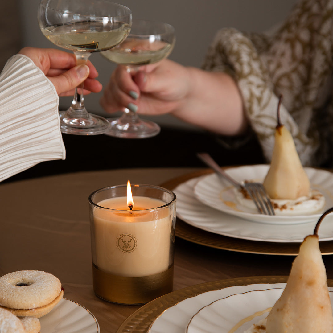Two people toast over poached pears and cookies as the Votivo Gilded Pear 10 oz. Holiday Candle glows on the table, creating a warm and festive scene.