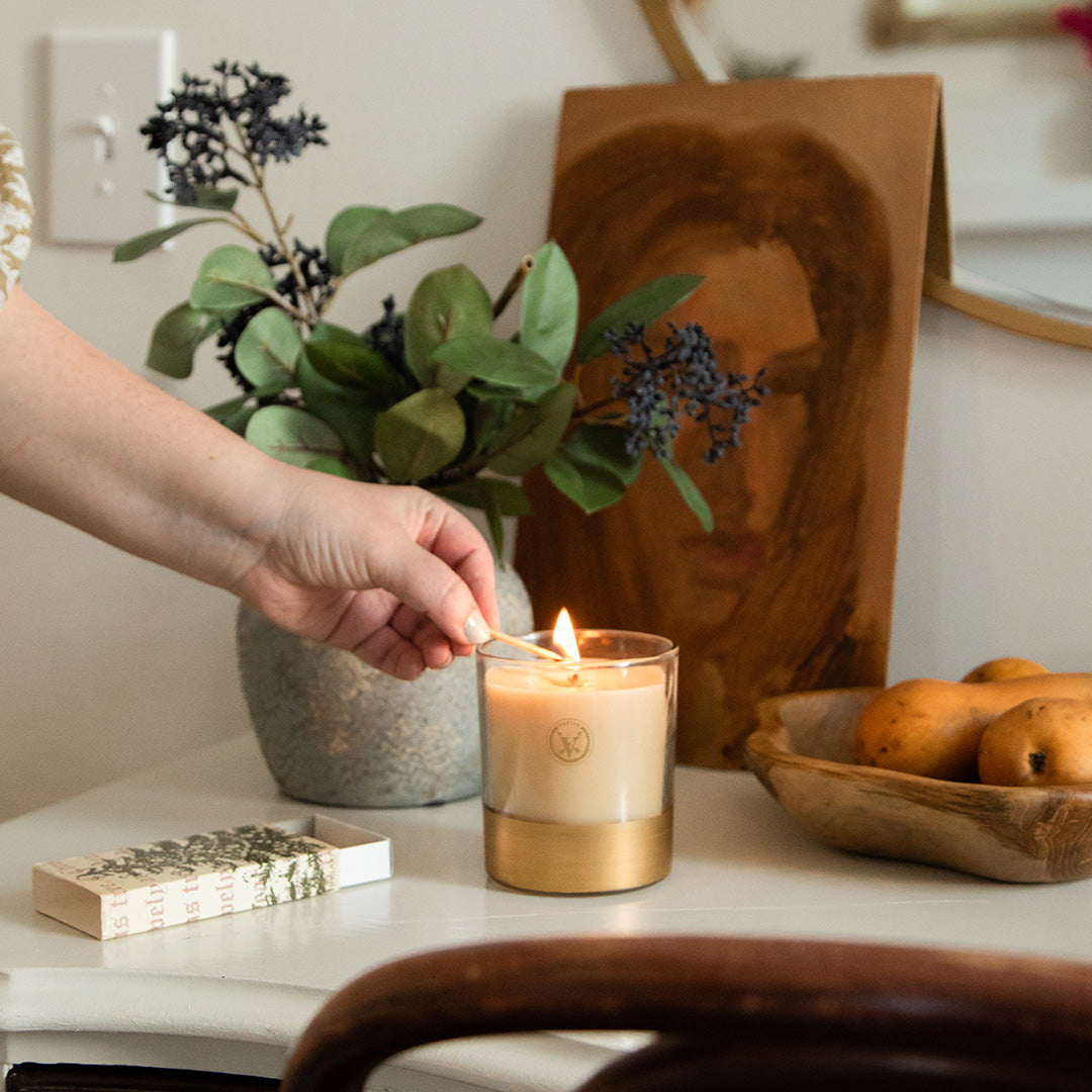 A hand lights the Votivo Gilded Pear 10 oz. Holiday Candle with a match on a table set with greenery in a vase, a wooden bowl of gilded pears, a matchbox, and a painting of a womans face in the background.