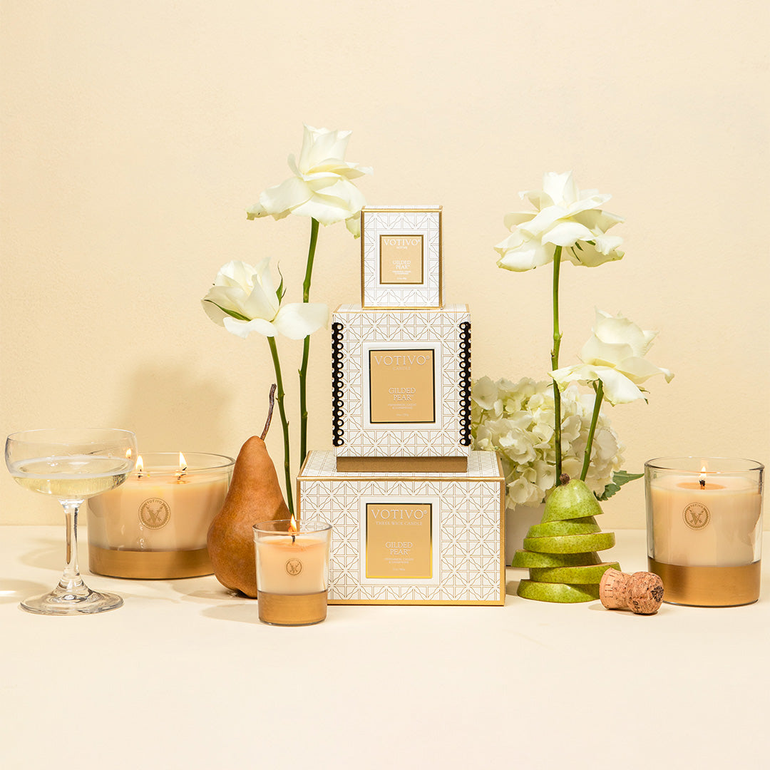 A display of Votivo’s Gilded Pear 10 oz. Holiday Candle with its box, set among white roses, a whole pear and slices, a champagne coupe, and cork on a cream surface—featuring luxurious soy blend wax.