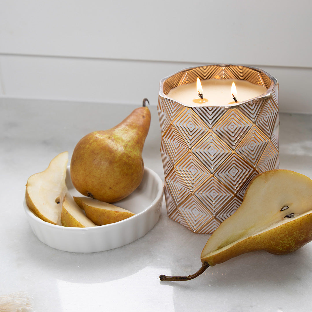 The Votivo Gilded Pear Holiday Decorative Candle, made from a soy blend wax, sits in a stylish vessel on marble beside fresh pears and adds warmth and elegance to any space.