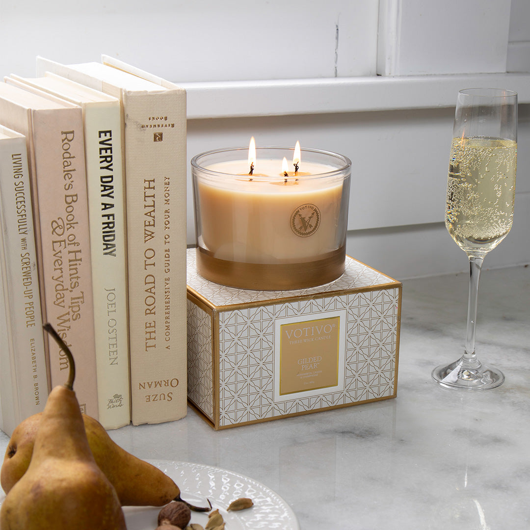 The Votivo Gilded Pear Holiday 3 Wick Candle sits lit on a decorative box next to champagne, cream books, and pears atop a white marble surface.