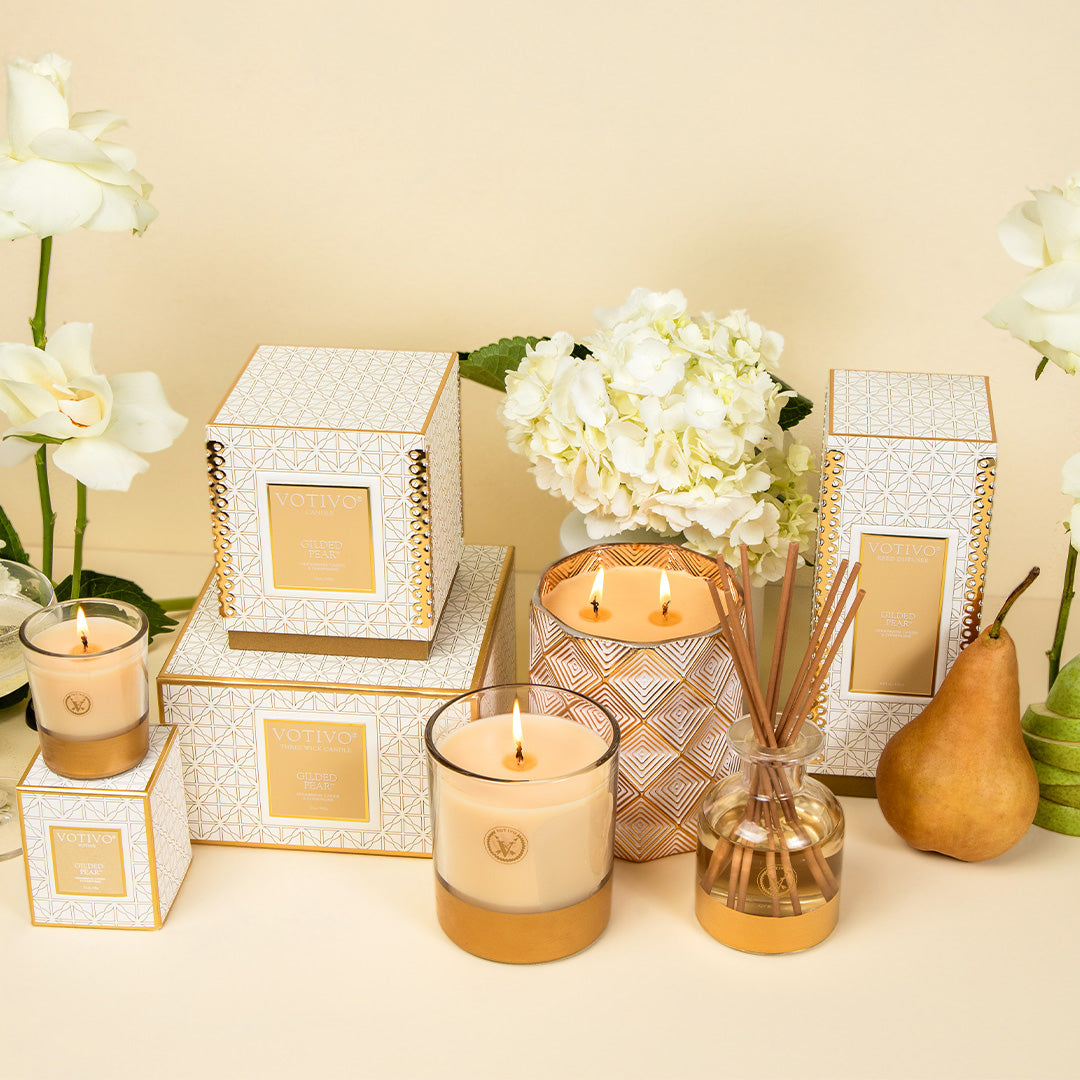 A display of Votivos Gilded Pear 10 oz. Holiday Candle, coordinating reed diffuser, and white and gold geometric boxes is arranged among white roses and hydrangeas on a beige background.