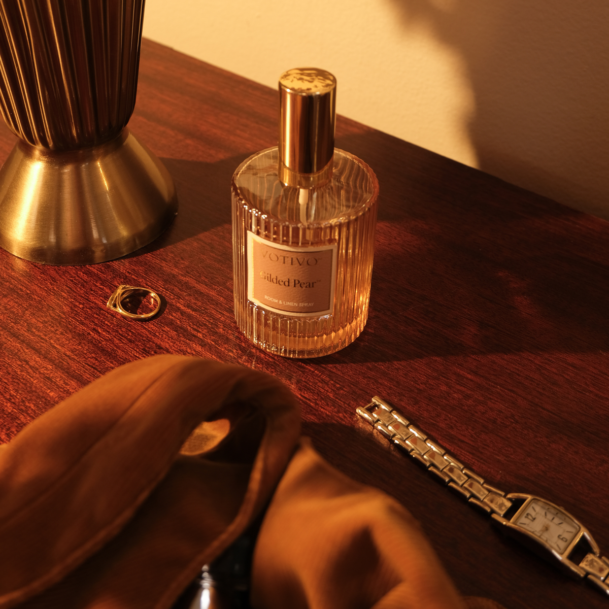 A bottle of Gilded Pear Room & Linen Spray sitting on a wood nightstand with jewelry around it