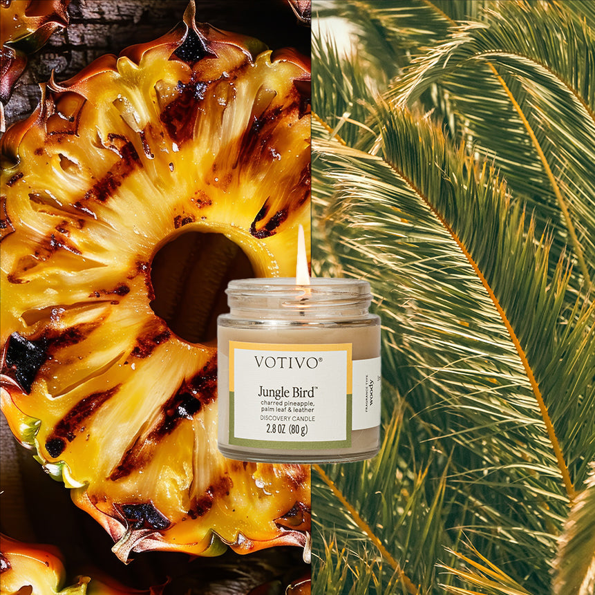 A Votivo Jungle Bird Discovery Candle sits between juicy grilled pineapple slices and green palm leaves, creating a vibrant, tropical scene with a long-lasting fragrance.