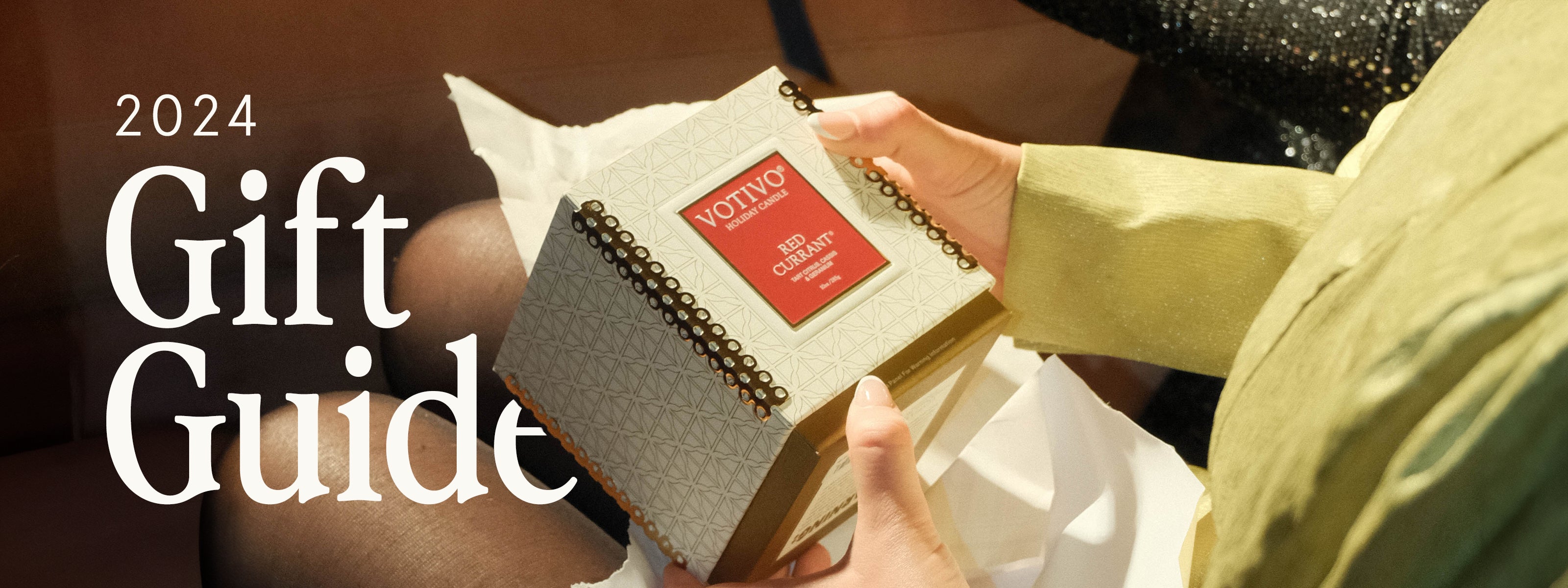 A person in a green sleeve holds a decorative box with a Votivo Red Currant candle. The text reads “2024 Gift Guide” in large white letters.