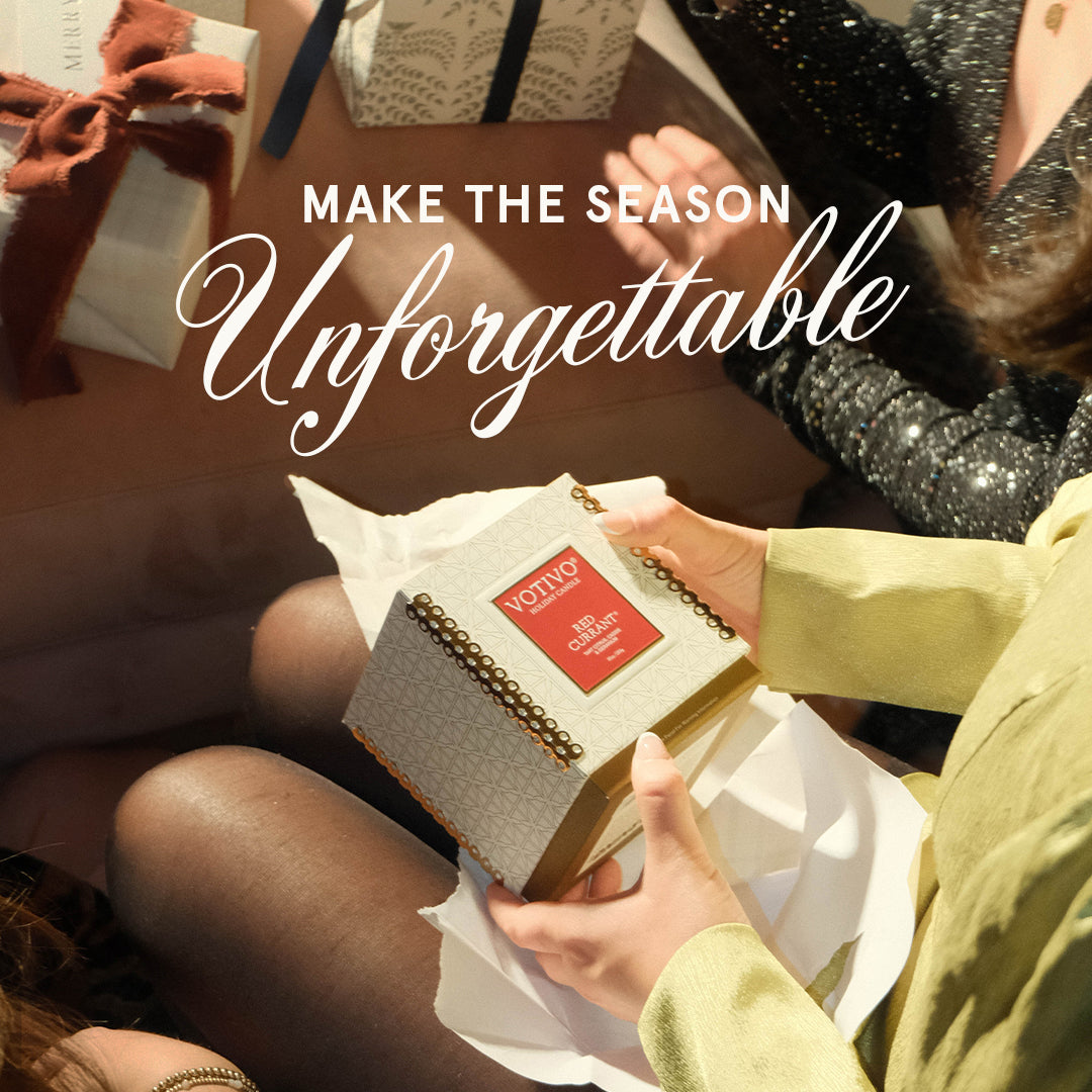 Three people in festive attire exchange gifts. One holds a Votivo Red Currant 10 oz. Holiday Candle. The words “Make the Season Unforgettable” appear above the scene.
