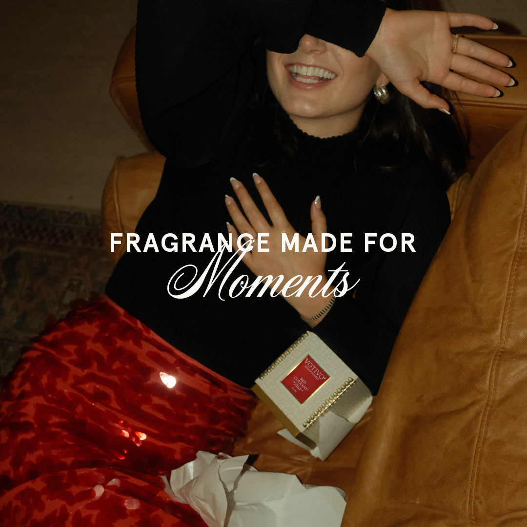 A smiling woman in a black top and red skirt leans on a brown leather couch, playfully covering her face. Nearby is the Votivo Red Currant 10 oz. Holiday Candle. A perfume box is also visible with text: “FRAGRANCE MADE FOR Moments.”.