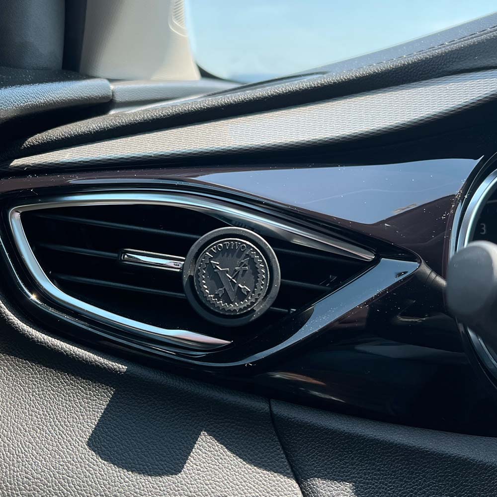 Close-up of a car air vent with a round, black Votivo Clean Crisp White Car Fragrance (2 Pack) attached to the grille. Sunlight streams in, illuminating the dashboard as the fresh scent enhances your drive.