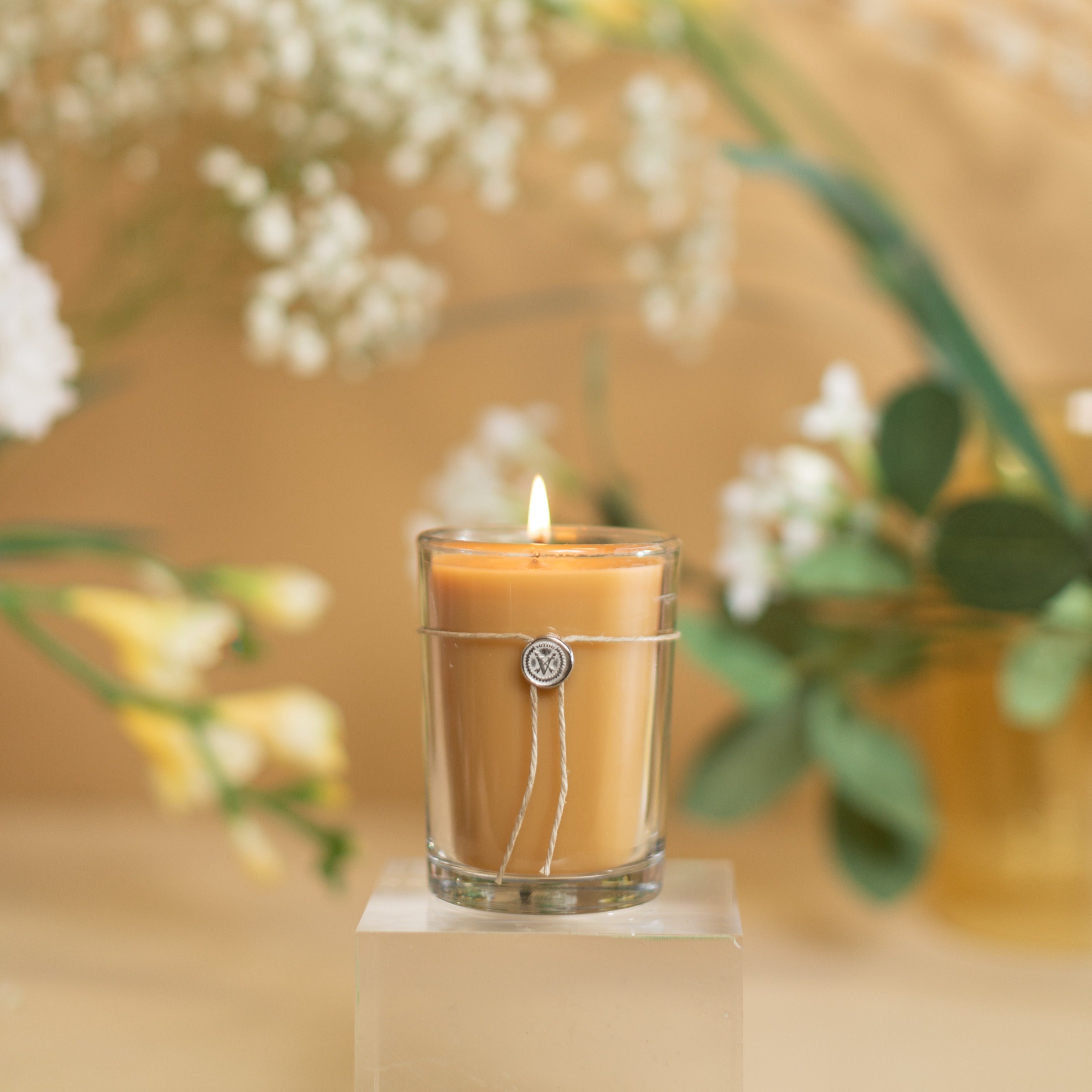 The Votivo Honeysuckle 6.8 oz. Aromatic Candle, crafted from a soy blend and displayed in a glass holder with a charm and string, sits on a clear pedestal with soft-focus white and yellow honeysuckle flowers behind it.