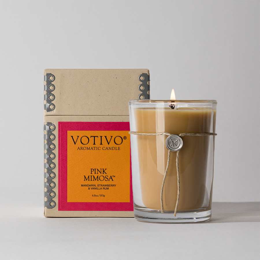 A Votivo Pink Mimosa 6.8 oz. Aromatic Candle in a glass jar with twine and a wax seal sits before its decorative box, which displays a red and orange label with scent details. The candle is made from a soy blend wax.