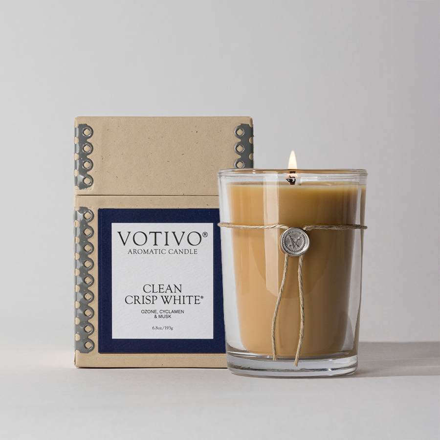 A lit Votivo Clean Crisp White 6.8 oz. Aromatic Candle glows in a clear glass jar with a string and charm, set before its beige and blue box. Crafted from soy blend wax, it stands out elegantly against a light background.