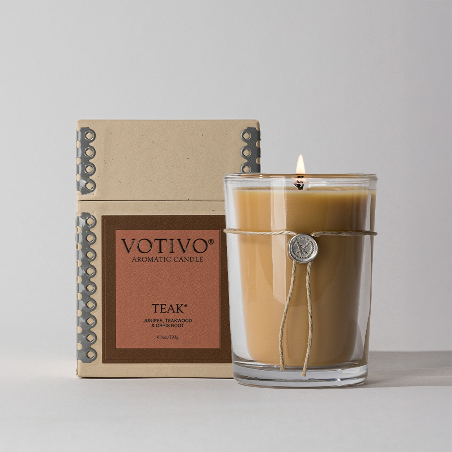 A brown Votivo Teak 6.8 oz. Aromatic Candle made from soy blend wax sits in a glass holder with a string and charm, placed before its box, evoking the rich scent of teakwood.