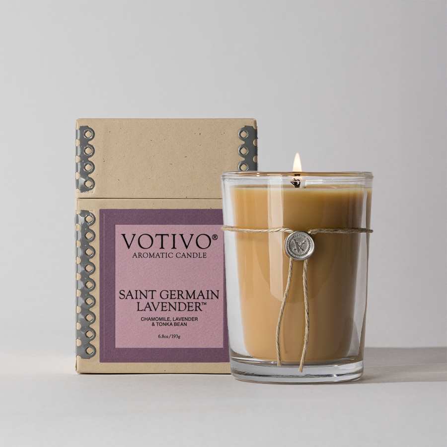 A lit Votivo Saint Germain Lavender 6.8 oz. aromatic candle, made from soy blend wax, sits in a clear glass holder with twine and a wax seal, displayed in front of its decorative purple and beige box.