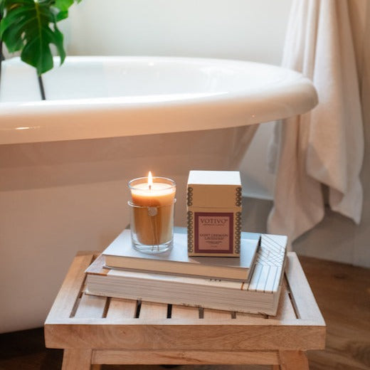 A Votivo Saint Germain Lavender 6.8 oz. Aromatic Candle sits, boxed and unboxed, on books atop a wooden stool beside a white bathtub in a cozy bathroom, with a plant and towel visible in the background.