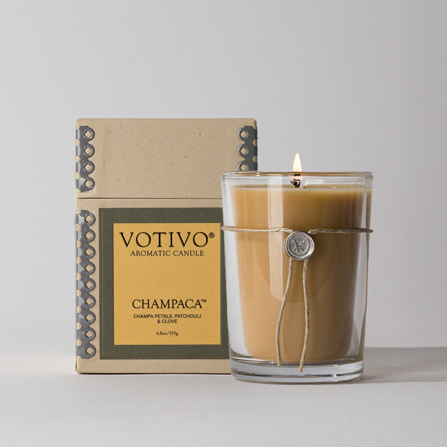 The Votivo Champaca 6.8 oz. Aromatic Candle, made with soy blend wax and wrapped in twine with a seal, sits before its beige and yellow box and features scent notes of champa petal, patchouli, and clove.