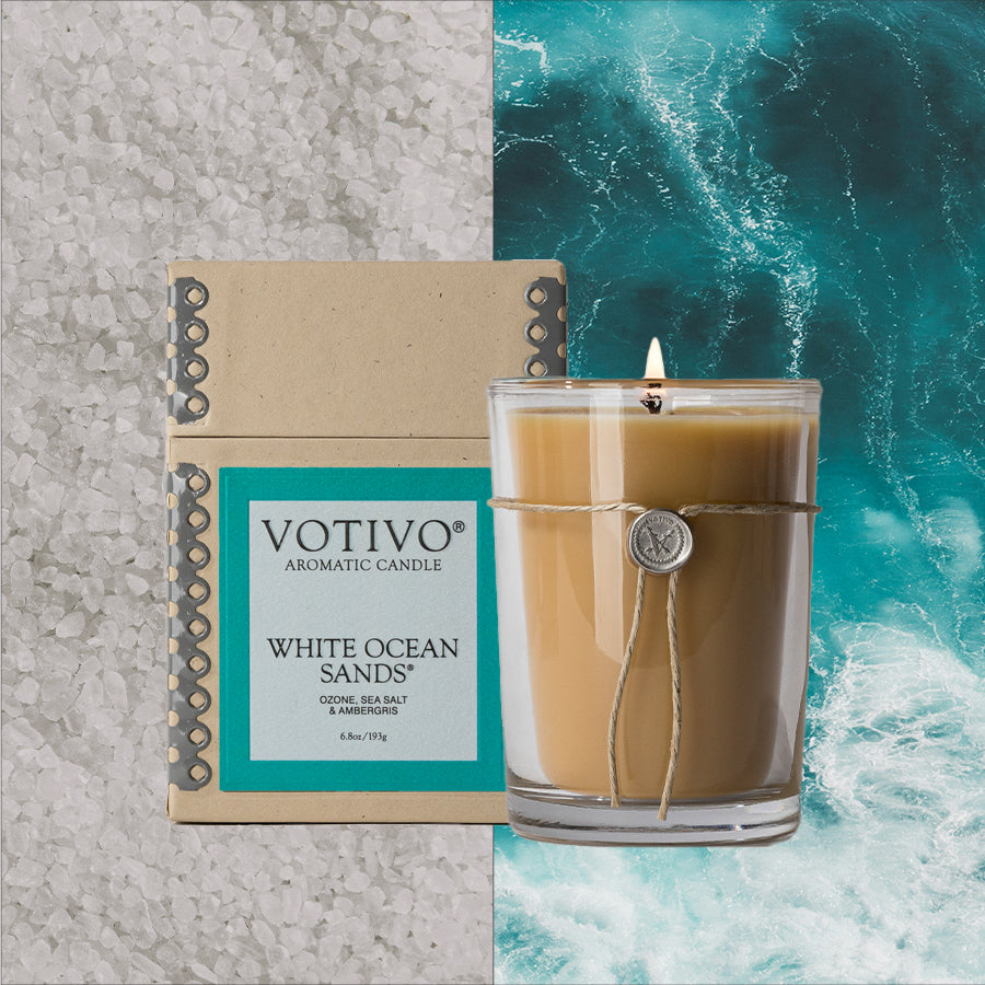 The Votivo White Ocean Sands 6.8 oz. Aromatic Candle, crafted with a soy blend wax and displayed in a glass jar next to its box, rests amid coarse sea salt crystals and blue ocean waves.