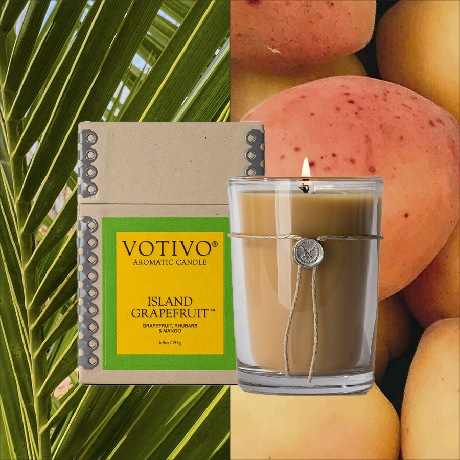 A Votivo Island Grapefruit 6.8 oz. Aromatic Candle in a glass jar rests next to its box amid palm leaves and ripe mangoes, highlighting the soy blend wax and vibrant fragrance.