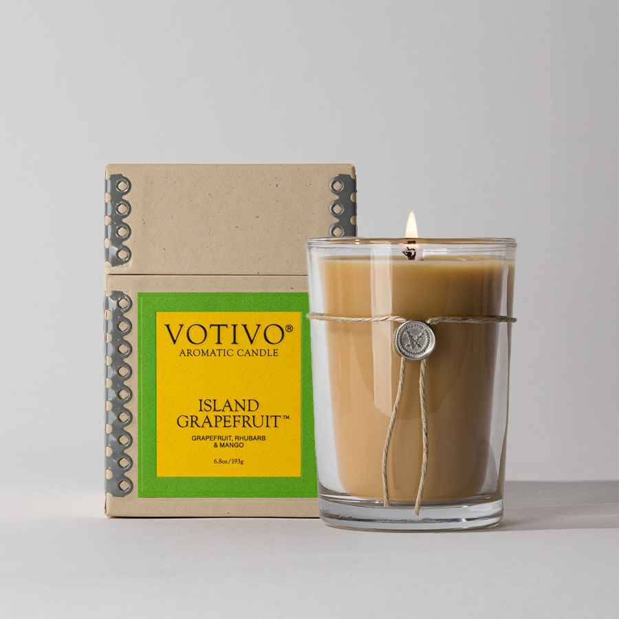 The Votivo Island Grapefruit 6.8 oz. Aromatic Candle, made with a soy blend and presented in a twine-wrapped glass jar, sits beside its yellow and green box on a light gray surface.