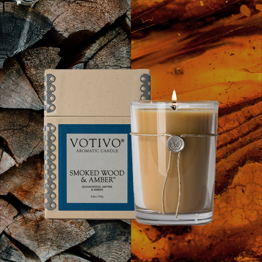 The Votivo Smoked Wood & Amber 6.8 oz. Aromatic Candle, made with a soy blend and housed in a glass jar, is shown beside its box with wood to the left and glowing amber hues to the right.