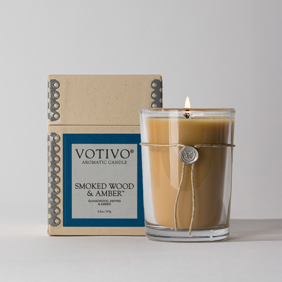 A lit Votivo Smoked Wood & Amber 6.8 oz. aromatic candle made from soy blend wax sits in a glass jar with a decorative seal and twine, next to its packaging, all set against a neutral background.