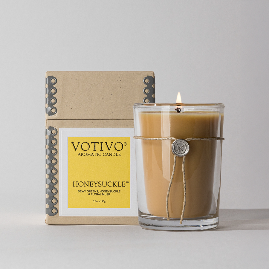 The Votivo Honeysuckle 6.8 oz. Aromatic Candle, crafted with a soy blend wax in a clear glass jar with decorative string and seal, glows warmly in front of its yellow-labeled box.