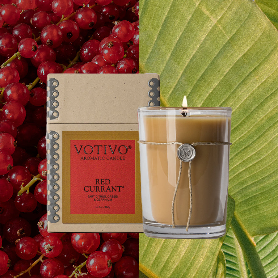 A Votivo Red Currant 6.8 oz. Aromatic Candle and its box sit beside a lit soy blend candle, set against fresh red currants and a green leaf, capturing the signature fragrance of Votivo’s Red Currant candle.