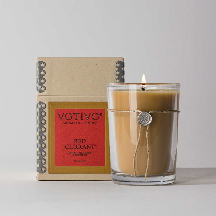 A lit Votivo Red Currant 6.8 oz. Aromatic Candle glows in a clear glass jar, tied with string and seal, placed before its decorative box on a light gray background.