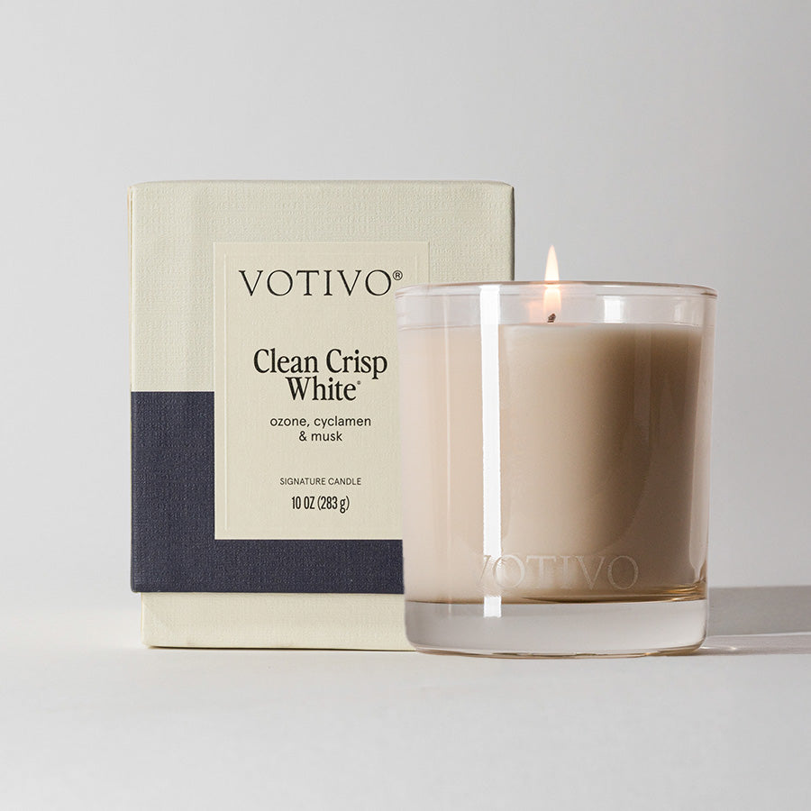 A Votivo Clean Crisp White 10 oz. Signature Candle made from a soy blend sits in a clear glass jar before its beige and black box, offering notes of ozone, cyclamen, and musk for a unique fragrance experience.