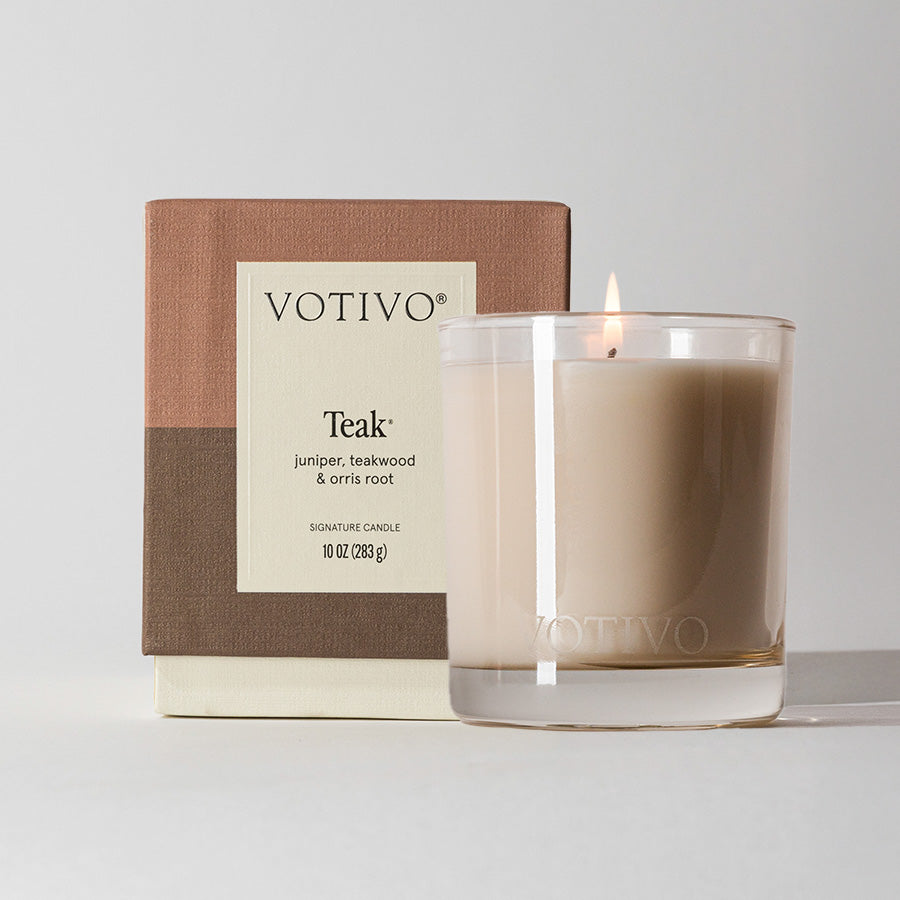 A lit Votivo Teak 10 oz. Signature Candle in a clear glass holder sits before its box, labeled with notes of juniper, teakwood, and orris root. This woody candle features earthy brown tones and a soy blend wax formula.