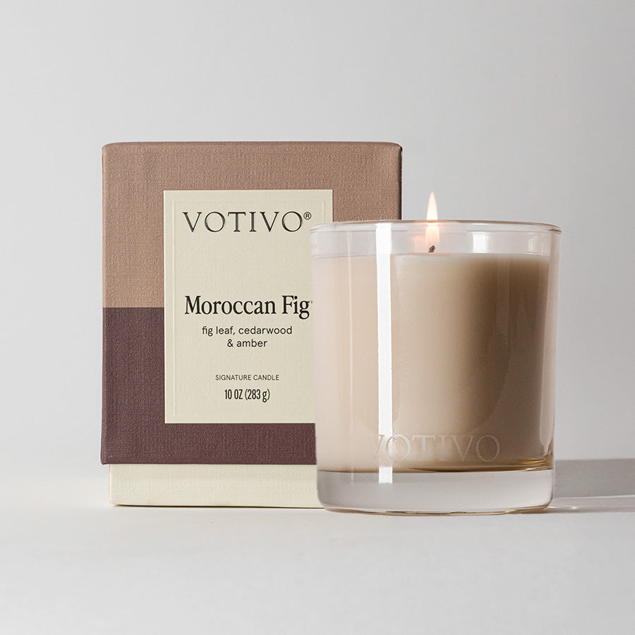 A lit Votivo Moroccan Fig 10 oz. Signature Candle made from a soy blend rests in a clear glass holder, set before its beige and brown box featuring scent notes and product details.