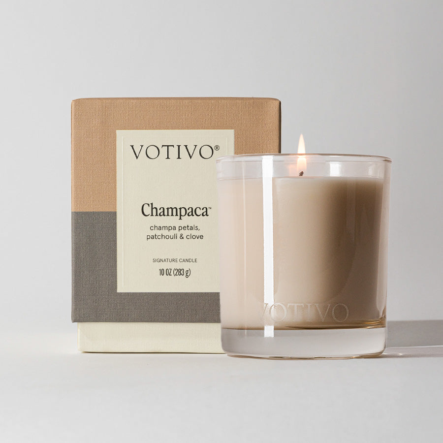 A lit Votivo Champaca 10 oz. Signature Candle in a clear glass holder sits by its beige and grey box, featuring scent notes of champa petals, patchouli, and clove in a unique soy blend fragrance.