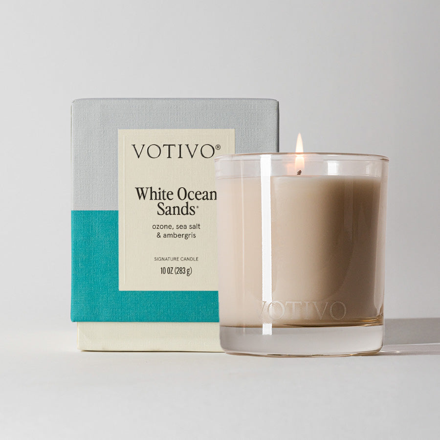 The Votivo White Ocean Sands 10 oz. Signature Candle, made from a soy blend, glows in a clear glass holder and features notes of ozone, sea salt, and ambergris; its box displays a teal and light gray design.