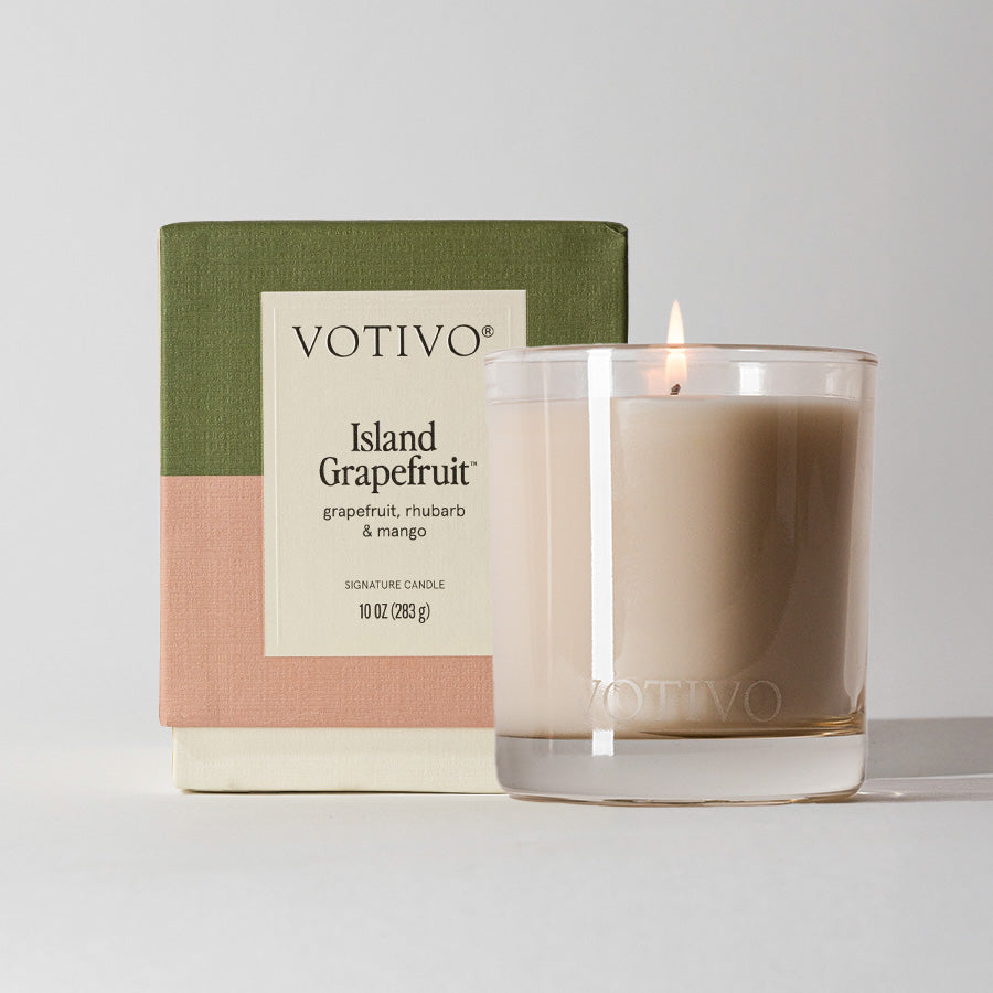 A lit Votivo Island Grapefruit 10 oz. Signature Candle, crafted from soy blend wax, sits in a clear glass holder in front of its green, pink, and beige color-blocked box featuring product details on the front.