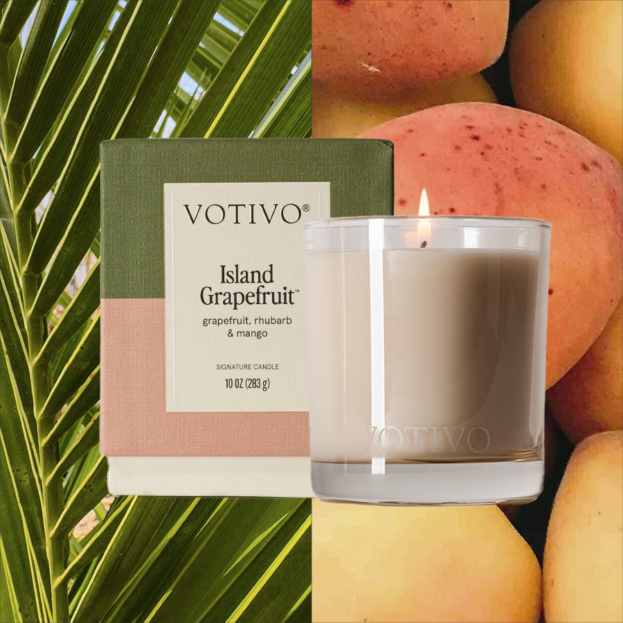 The Votivo Island Grapefruit 10 oz. Signature Candle, made with a soy blend wax and presented in a glass jar, sits next to its box alongside a palm leaf and mangoes, capturing lively tropical and fruity aromas.