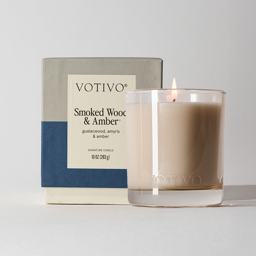 A lit Votivo Smoked Wood & Amber 10 oz. Signature Candle in a clear glass holder rests by its box, featuring notes of guaiacwood, amyris, and amber. Crafted from soy blend wax, it sits on a light surface against a plain background.