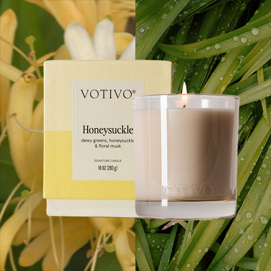 A Votivo Honeysuckle 10 oz. Signature Candle sits beside its yellow-and-white box, with honeysuckle flowers and dew-covered green leaves in the background. Made from premium soy blend wax.