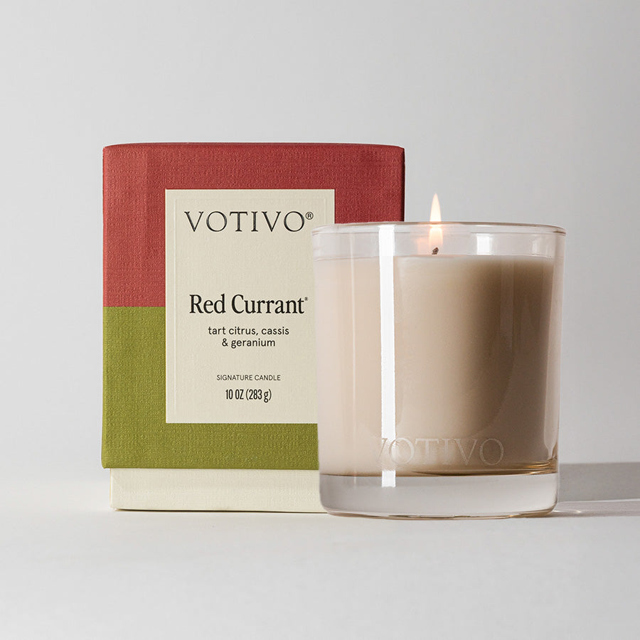 A lit Votivo Red Currant 10 oz. Signature Candle, crafted with soy blend wax, sits in a clear glass holder beside its red and green box labeled Red Currant and scent notes: tart citrus, cassis & geranium.