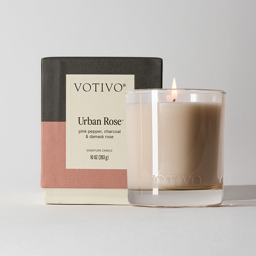 A lit beige Votivo Urban Rose 10 oz. Signature Candle made with soy blend wax sits before its box, which features stylish color blocks and scent notes of pink pepper, charcoal, and damask rose.
