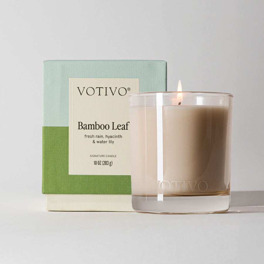 A lit Votivo Bamboo Leaf 10 oz. Signature Candle, made with a soy blend wax in a clear glass holder, is set before its green and blue box featuring fresh rain, hyacinth, and water lily fragrance notes.