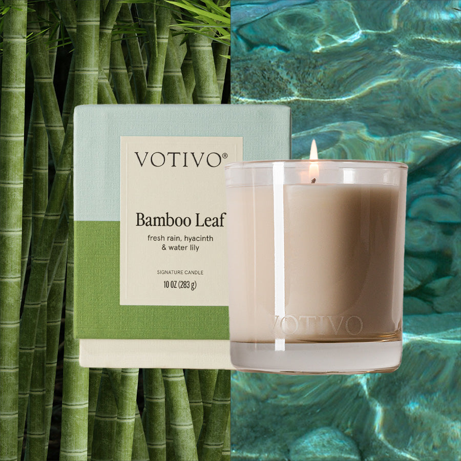 The Votivo Bamboo Leaf 10 oz. Signature Candle is displayed in a clear glass jar next to its green and white box, set between bamboo stalks and sparkling water. The soy blend wax offers a clean, long-lasting burn.