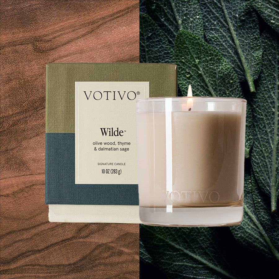A lit Votivo Wilde 10 oz. Signature Candle made with soy blend wax sits beside its box on a wood-grain surface, surrounded by green leaves, releasing the unique fragrance of olive wood, thyme, and Dalmatian sage.