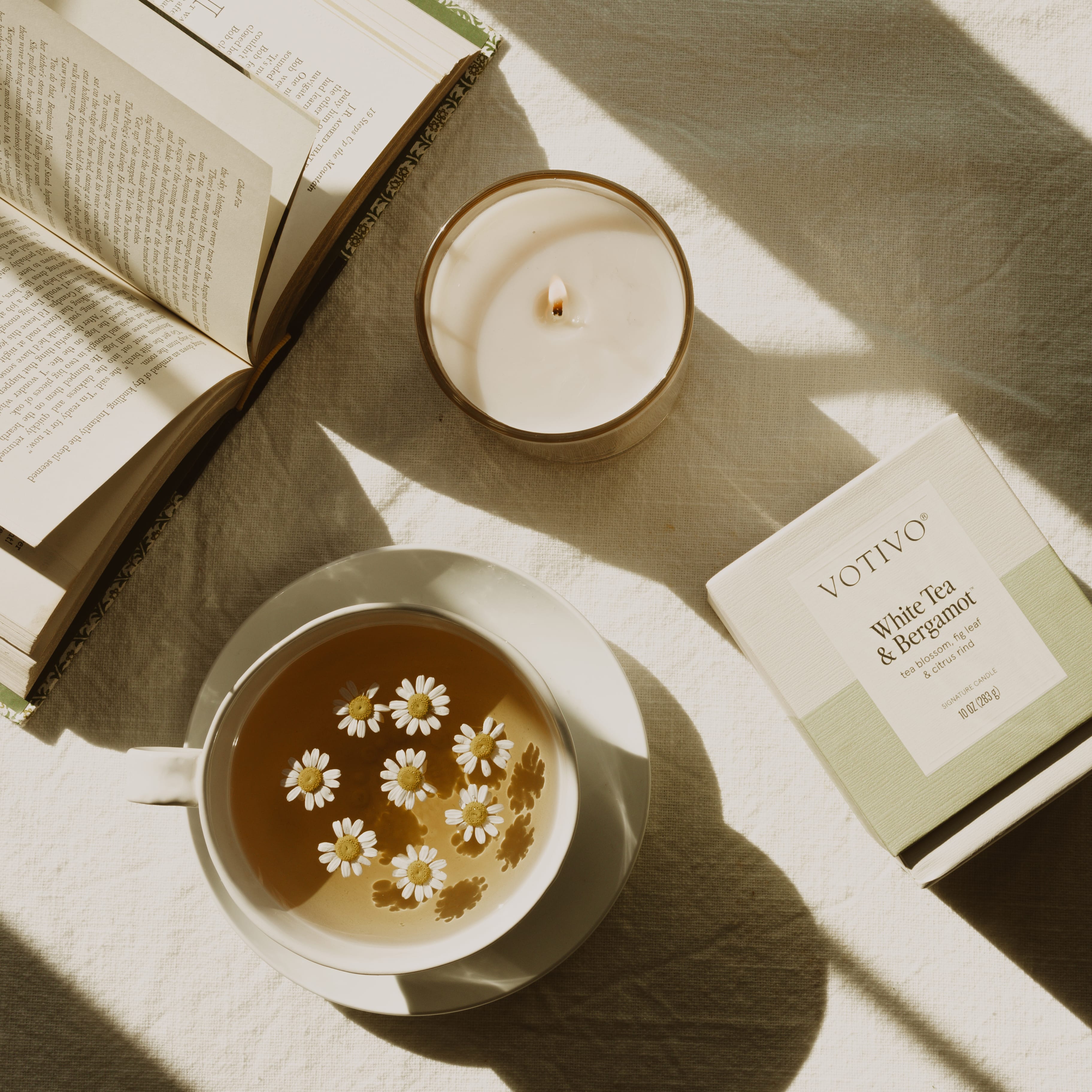 A cup of chamomile tea, an open book, a lit Votivo White Tea & Bergamot 10 oz. Signature Candle made with soy blend wax, and its box are arranged on a sunlit white cloth.
