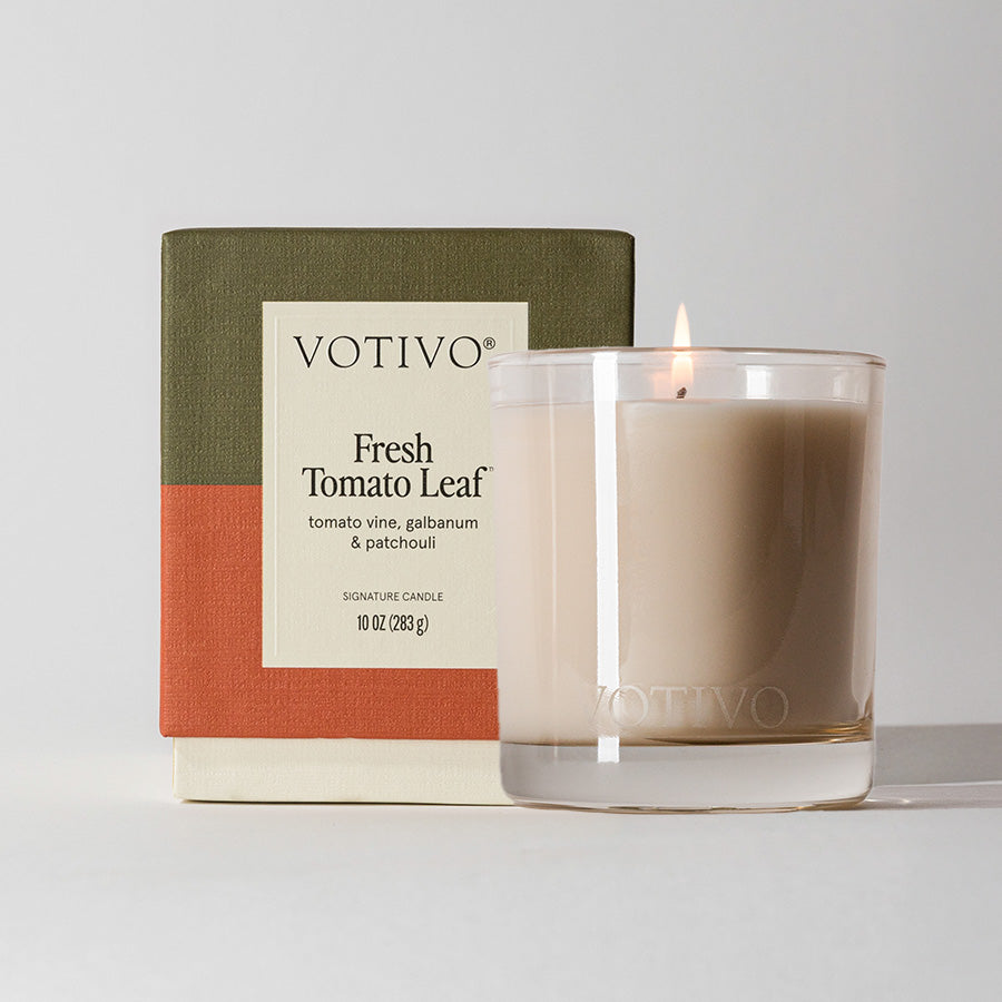 A lit Votivo Fresh Tomato Leaf 10 oz. Signature Candle in a clear glass holder sits before its box, featuring green, cream, and orange blocks. The soy blend wax fills the air with tomato vine, galbanum, and patchouli aromas.