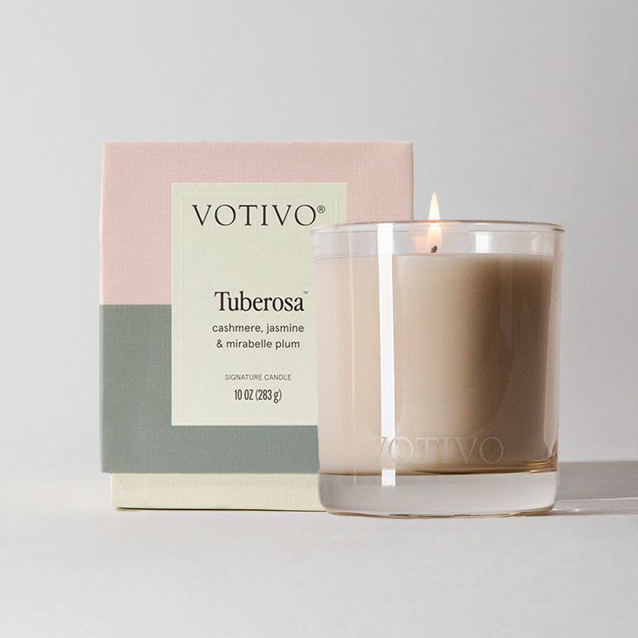 A lit Tuberosa 10 oz. Signature Candle by Votivo, made of soy blend wax and set in a clear glass holder, stands before a pastel box labeled with cashmere, jasmine & mirabelle plum fragrance notes.
